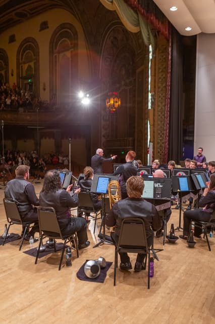 Brass band members applaud on stage as the conductor gestures, with audience in an ornate theatre.