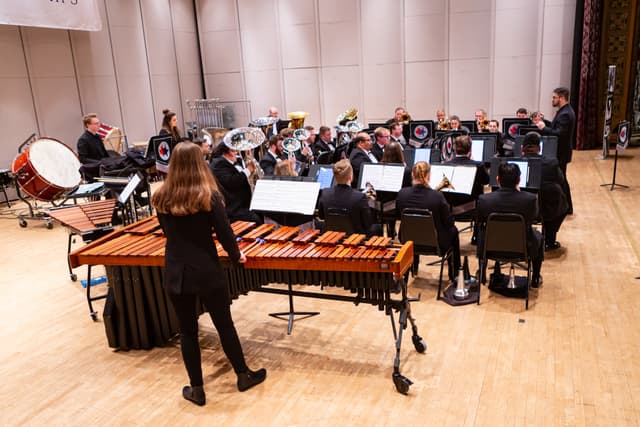 Brass band performing on stage with conductor, marimba and bass drum in foreground