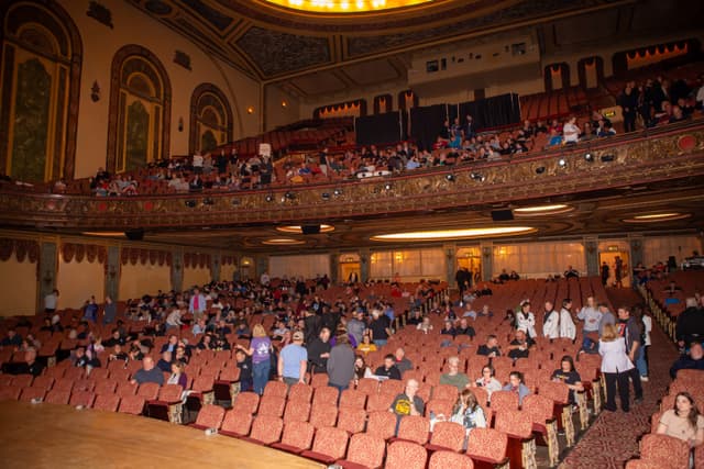 Spectators finding seats in an ornate historic theatre with gilded balcony, viewed from the stage