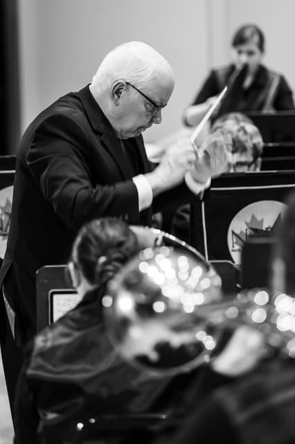 Conductor with raised baton leads a brass band, with blurred players and instrument bell in foreground
