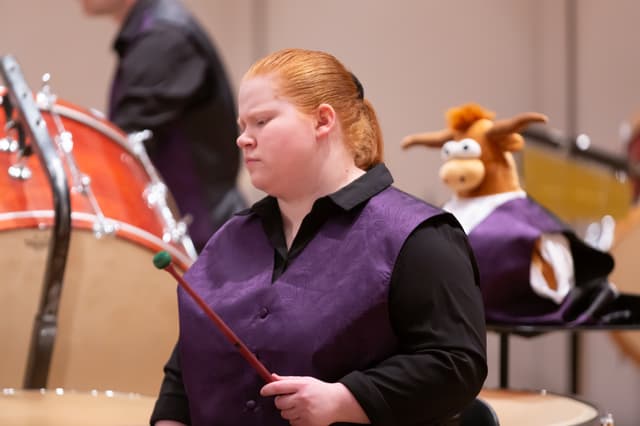 Percussionist holding a mallet beside drums, with a plush longhorn mascot in the background