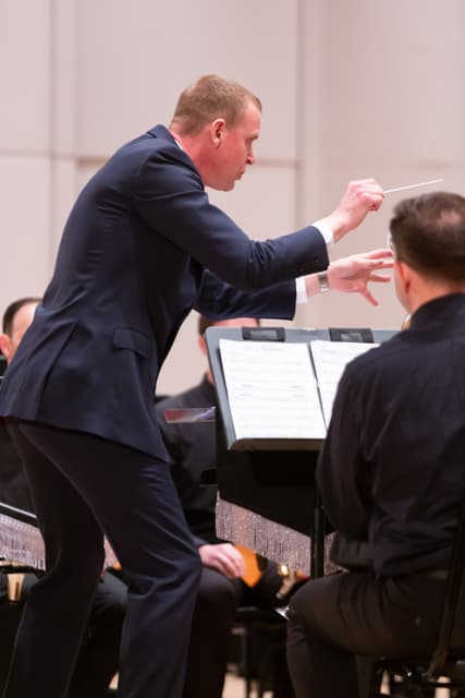 Conductor with baton leading seated brass band musicians in a concert hall