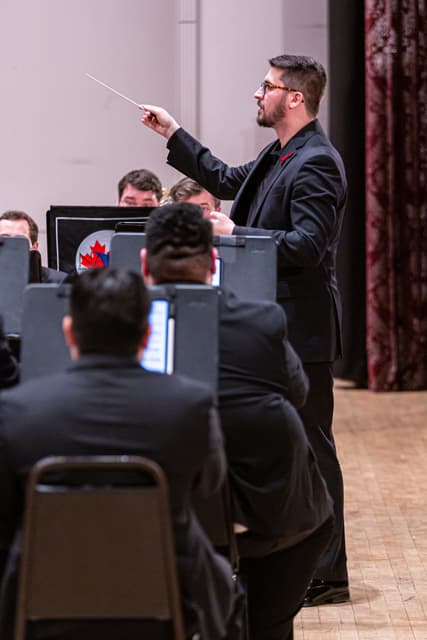 Conductor with raised baton leads seated band members on stage beside a red curtain