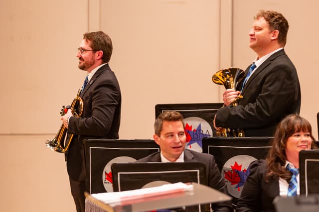 Brass band members on stage holding a flugelhorn and tenor horn beside NABBA music stand banners