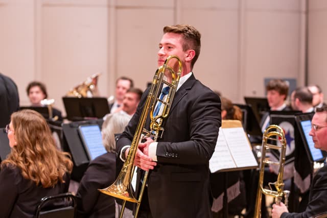 Standing trombonist in a dark suit holding his instrument, with seated brass band members behind him.