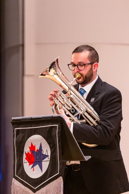 Euphonium player performing on stage behind a music stand with a NABBA banner