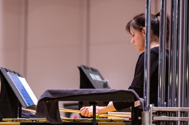 Percussionist holding mallets over a keyboard instrument beside tubular bells and music stands