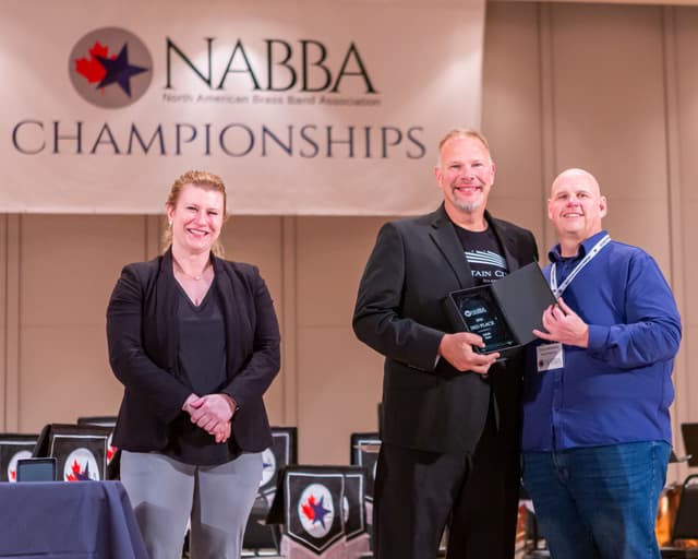 Award recipient holding a 3rd place Adult Solo plaque on stage with two smiling officials
