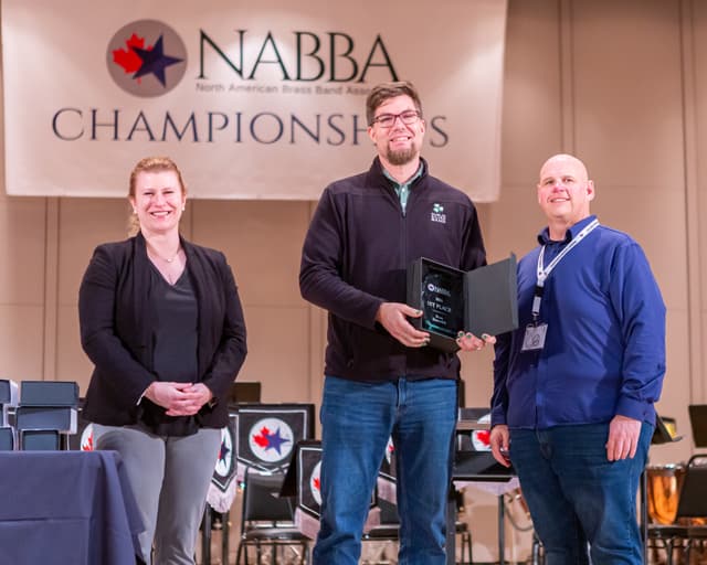 Three people on stage with a 1st place brass ensemble award plaque beneath the NABBA Championships banner