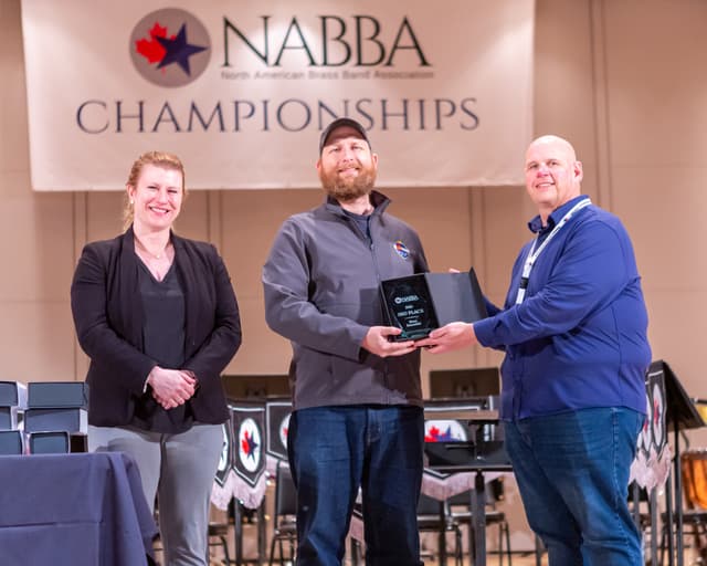 Three people on stage presenting a 3rd place Brass Ensemble award beneath a NABBA Championships banner