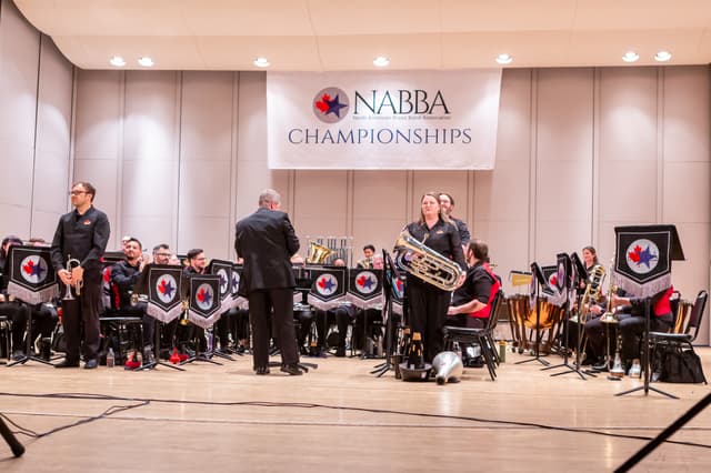 Brass band on stage under a NABBA Championships banner, with a cornet player, euphonium player and conductor standing.