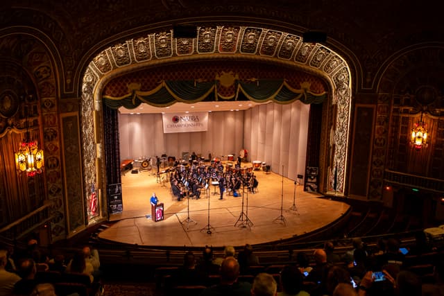 Brass band performing on a grand theatre stage under a NABBA Championships banner, viewed from the balcony