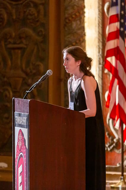 Speaker at a podium with microphone in an ornate theatre beside an American flag