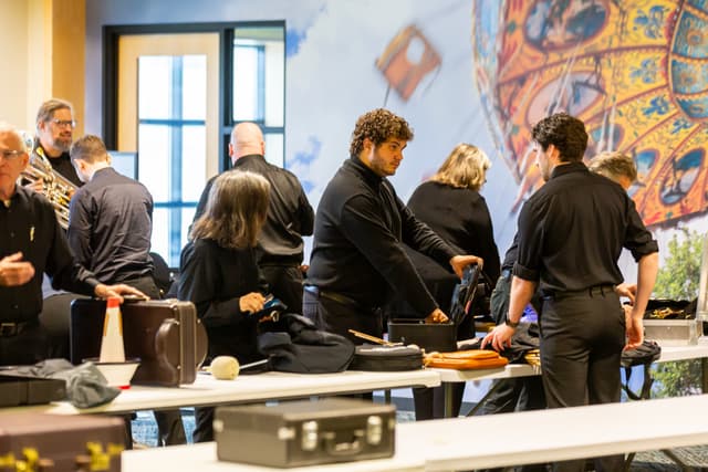 Musicians in black unpack instrument cases at tables in front of a colorful carousel mural.