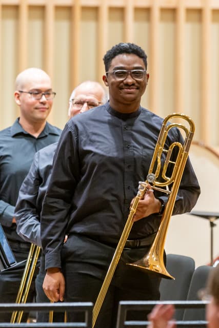 Smiling trombonist holding a trombone on stage with other brass players behind