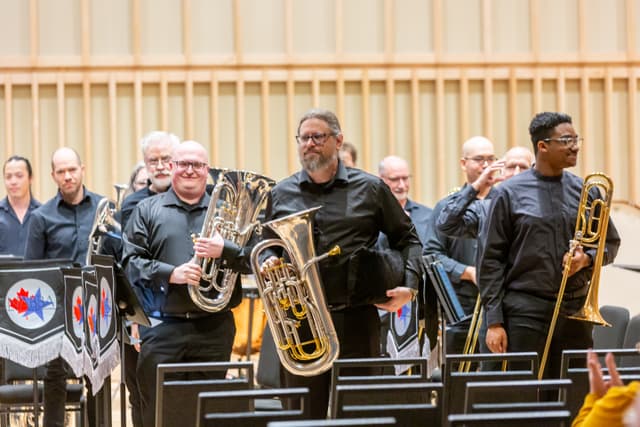 Band members in black carrying euphoniums and a trombone leave a wood-panelled stage past NABBA banners.