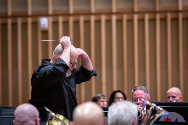 Conductor with raised baton leads a brass band in a wood-panelled hall