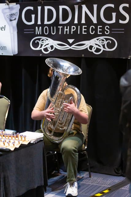 Seated musician playing a large silver tuba beneath a Giddings Mouthpieces banner at a trade stand