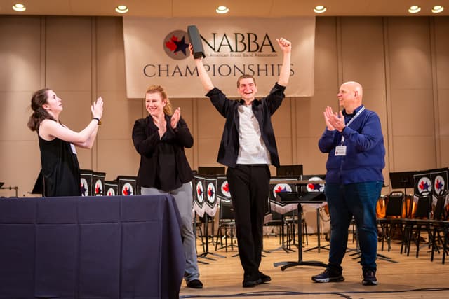 Smiling winner raising a trophy on stage as three people applaud at NABBA Championships
