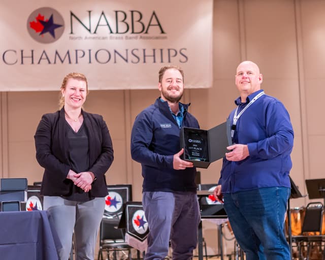 Three smiling people on stage holding a NABBA 2026 1st place Ten-Piece Ensemble award plaque