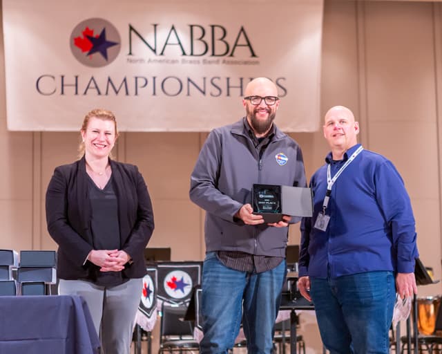 Three people on stage under a NABBA Championships banner, one holding a 2nd place glass award