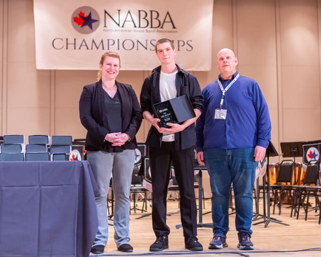 Three people on stage holding a 3rd place percussion ensemble award beneath a NABBA Championships banner