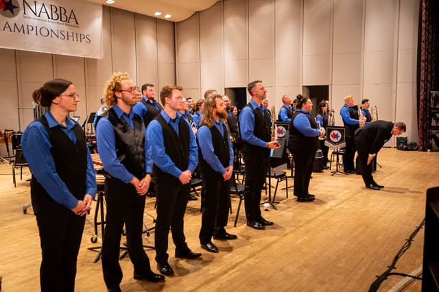 Brass band in blue shirts and black vests standing on stage as a conductor bows
