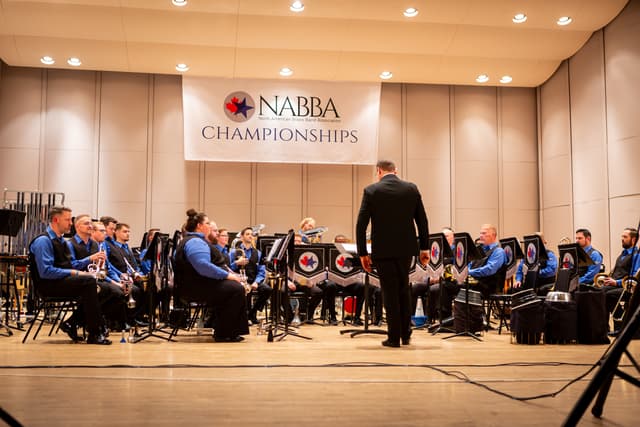 Conductor leading a seated brass band beneath a NABBA Championships banner on stage