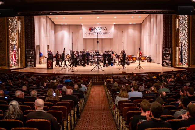 Brass band on a theatre stage under a NABBA Championships banner, with applauding audience in foreground
