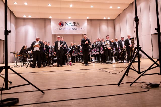 Brass band members and conductor standing on stage beneath a NABBA Championships banner