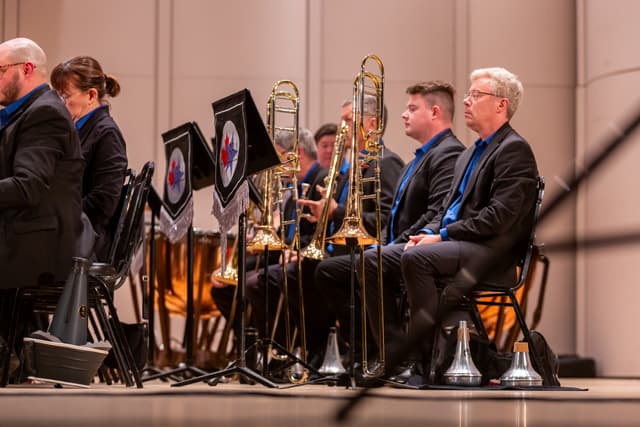 Seated trombone section in dark jackets and blue shirts beside banner-draped music stands on stage
