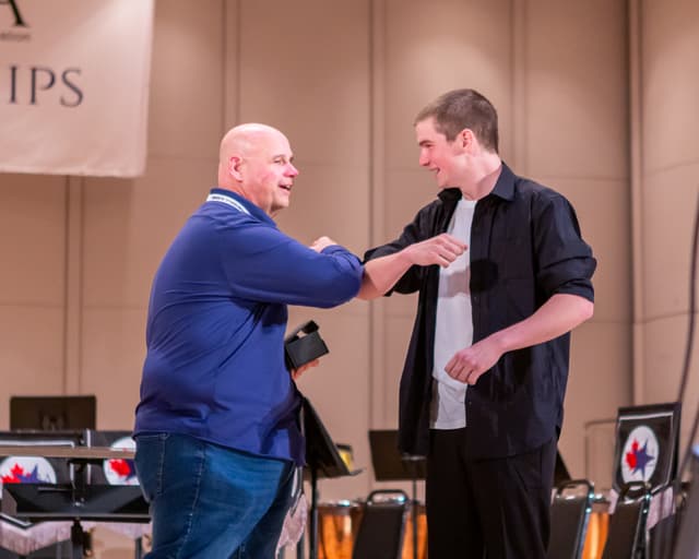 Two men bump elbows on stage at an awards presentation, one holding a small box.