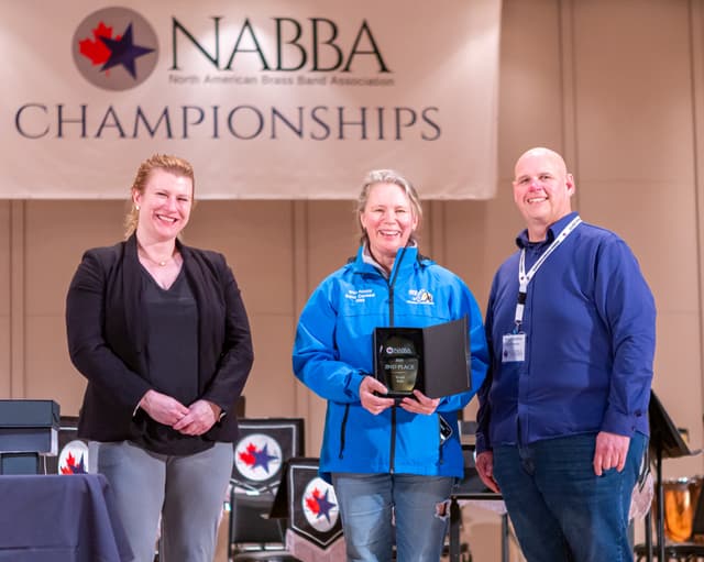Three smiling people on stage holding a 2nd place Youth Solo award under a NABBA banner