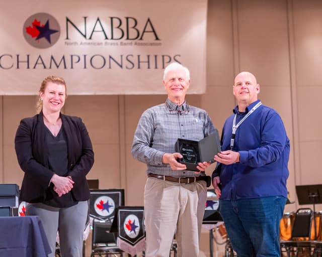 Three people on stage presenting a 2nd place Senior Solo award plaque beneath a NABBA Championships banner