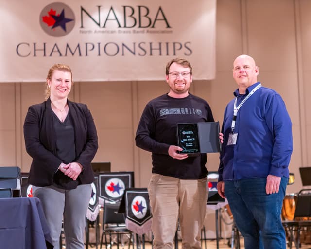 Three people on stage holding a 3rd place Ten-Piece Ensemble award beneath a NABBA Championships banner