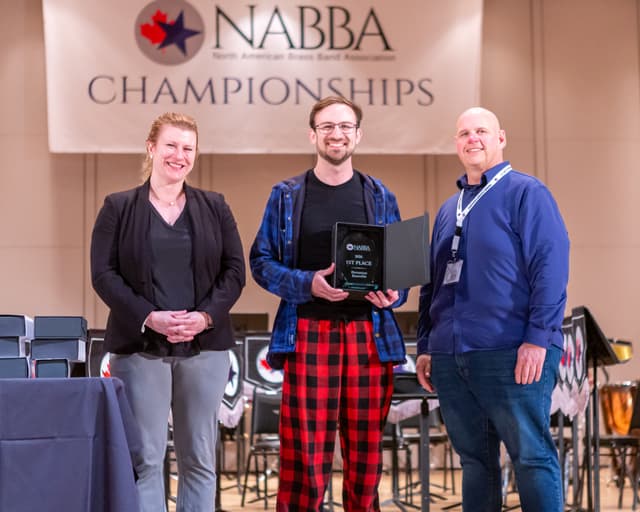 Three people on stage with a 1st Place Percussion Ensemble award plaque beneath a NABBA Championships banner