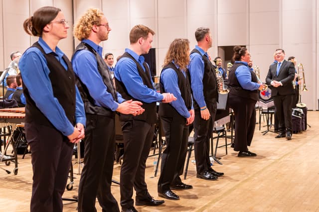 Brass band members in blue shirts and black vests standing in a line on stage
