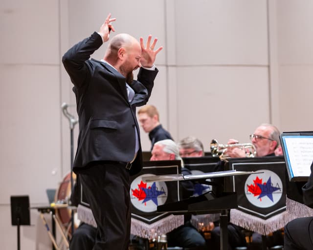 Conductor with raised hands leads a brass band on stage while a cornet player performs.
