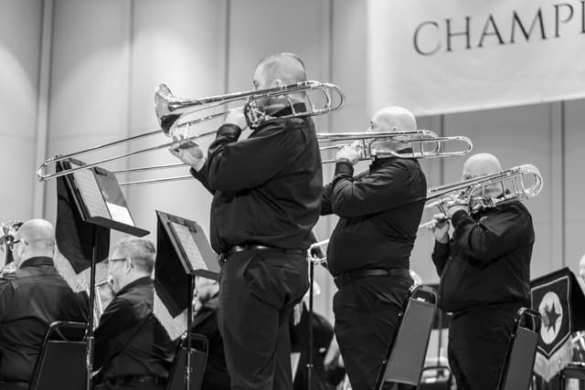 Three standing trombonists perform on stage with music stands and seated band members nearby.