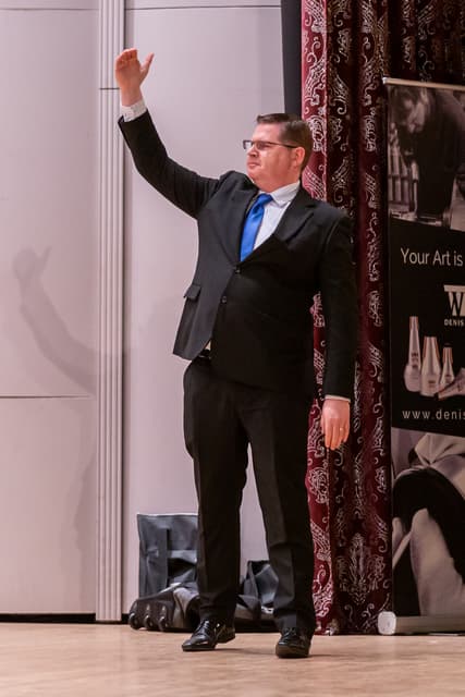 Conductor in dark suit raising one arm on stage beside curtain and banner