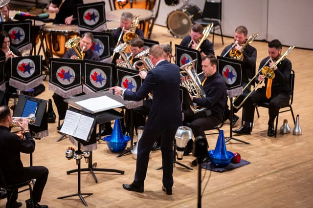 Conductor directing a brass band with trombones and euphoniums on stage, music stands bearing NABBA banners