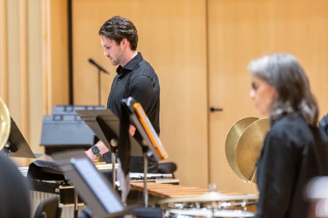 Percussionist at a marimba beside music stands and cymbals in a wood-panelled concert hall