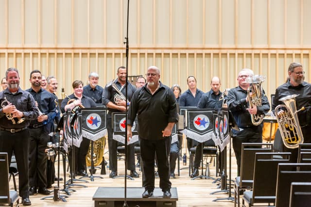 Conductor on podium with smiling brass band members standing behind holding instruments on a concert stage