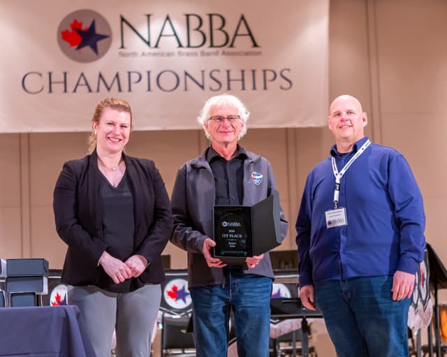 Three people on stage holding a 1st Place Senior Solo award plaque under a NABBA banner