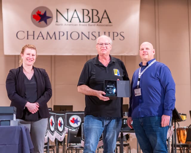 Three people on stage with a 3rd place Senior Solo glass award under the NABBA banner