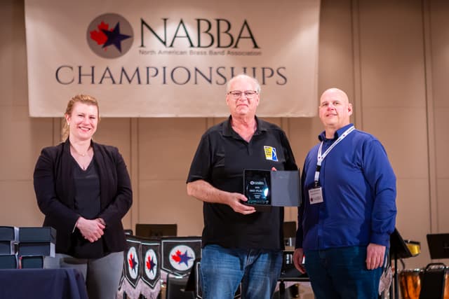 Three people on stage with a 3rd place Senior Solo award beneath a NABBA Championships banner