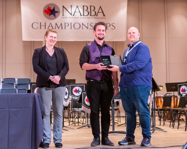 Three people on stage during an awards presentation at the NABBA Championships, holding a 2nd place plaque.