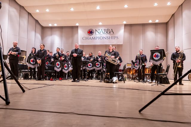 Brass band members standing on stage beneath a NABBA Championships banner, instruments in hand.