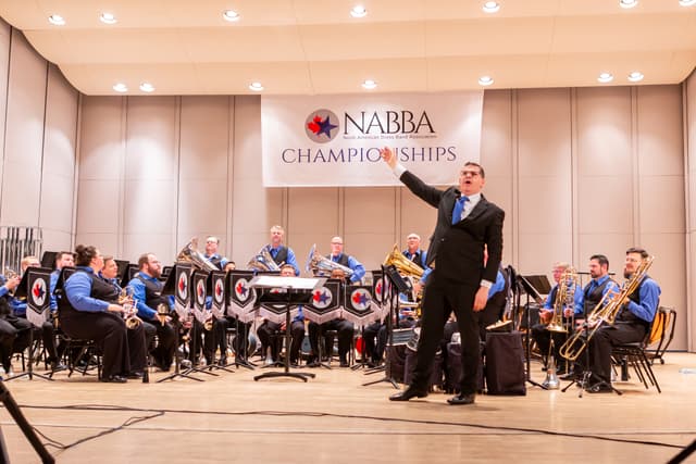 Conductor gesturing in front of a seated brass band beneath a NABBA Championships banner