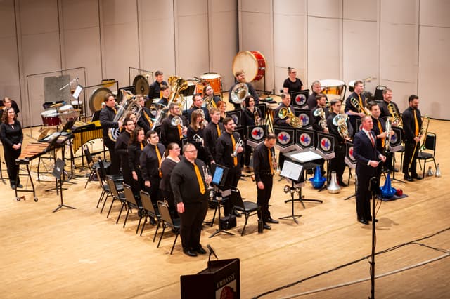 Brass band standing on stage with instruments and conductor, smiling at the audience after a performance